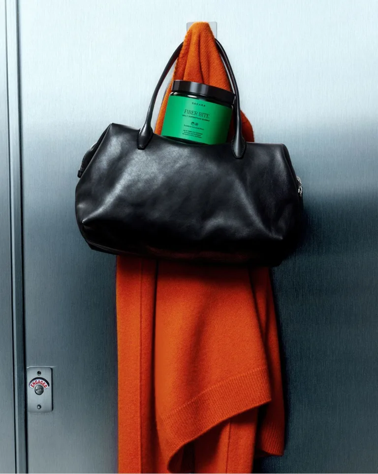 A sweater, bag, and Sakara Fiber Bite hanging from a hook on the back of a bathroom stall door.