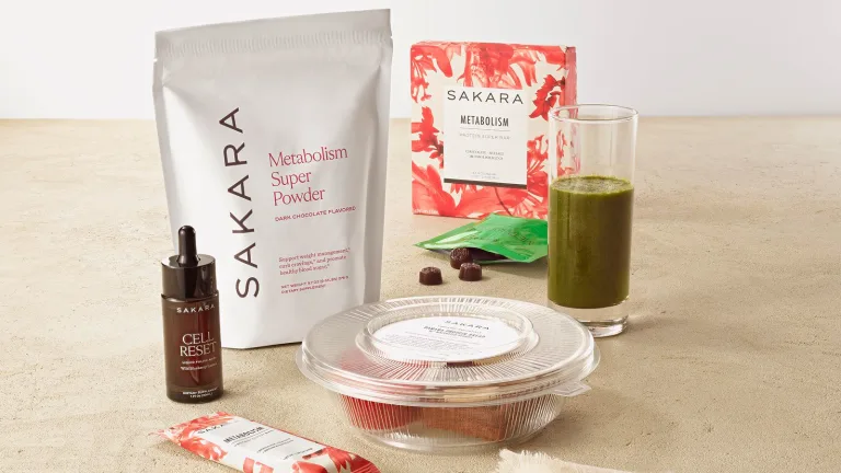 Assorted Sakara supplements (including Metabolism Super Powder, Metabolism Protein Super Bar, Cell Reset, and Metabolism Bite), a green smoothie, and packaged Sakara breakfast arranged on a neutral surface.